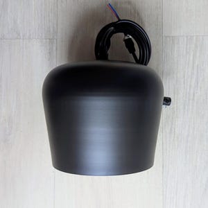 May include: A black pendant light fixture with a round, slightly curved shade. The fixture has a black wire cord with a plug.