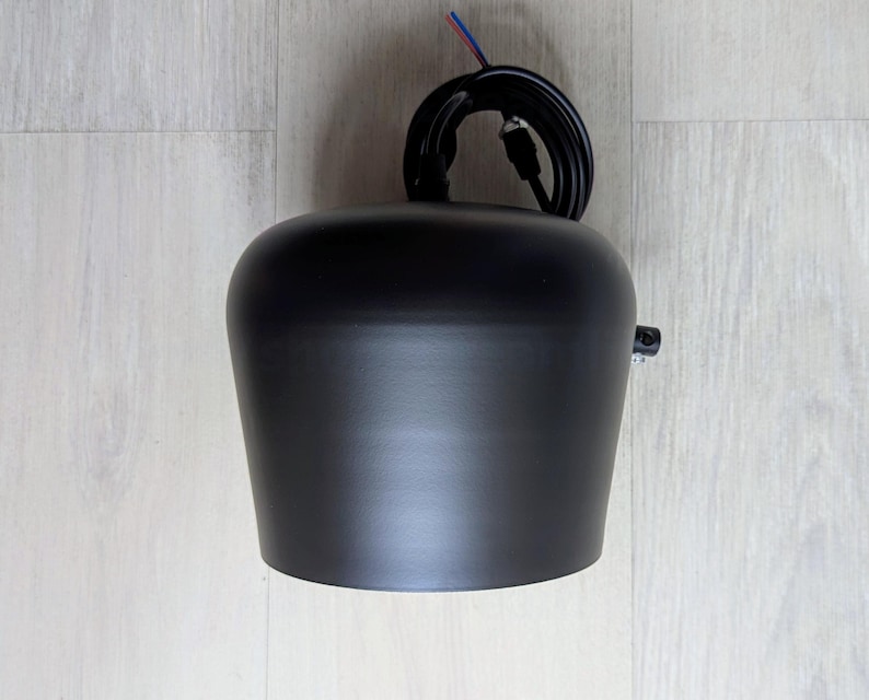 May include: A black pendant light fixture with a round, slightly curved shade. The fixture has a black wire cord with a plug.