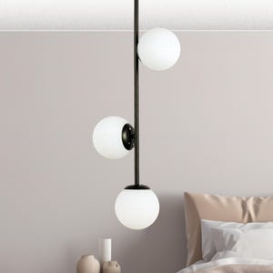 Op de afbeelding: Een moderne hanglamp met drie bolvormige witte glazen kappen. De kappen zijn gelijkmatig verdeeld over een zwarte metalen staaf. De lamp hangt aan het plafond, met een slaapkamer op de achtergrond.