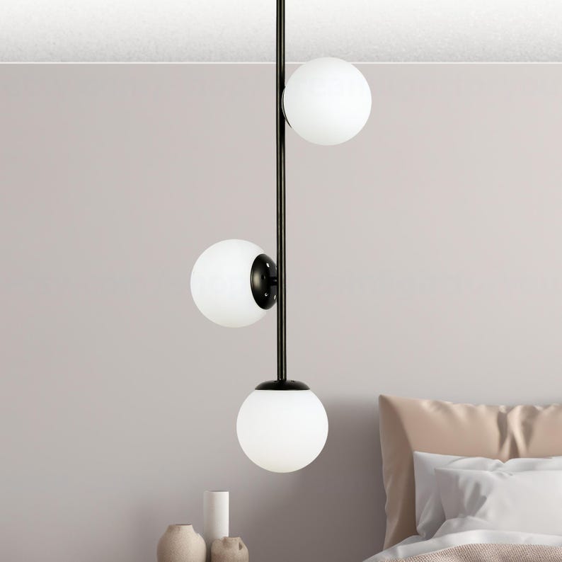 Op de afbeelding: Een moderne hanglamp met drie bolvormige witte glazen kappen. De kappen zijn gelijkmatig verdeeld over een zwarte metalen staaf. De lamp hangt aan het plafond, met een slaapkamer op de achtergrond.