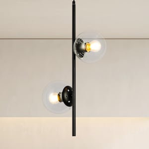 Op de afbeelding: Moderne zwarte hanglamp met twee heldere glazen bolvormige kappen. Elke bol bevat een warme, gloeiende gloeilamp. De armatuur hangt aan een zwarte stang, wat een minimalistisch en eigentijds ontwerp cre&euml;ert.