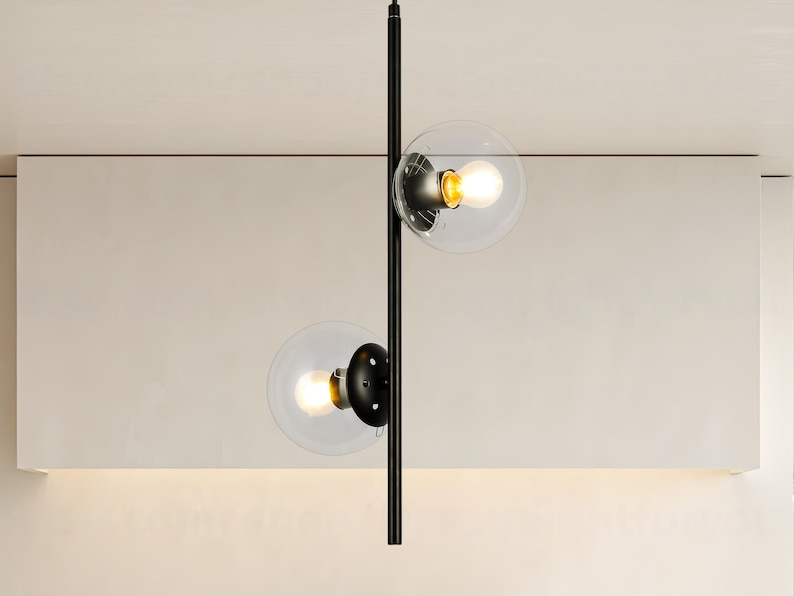 Op de afbeelding: Moderne zwarte hanglamp met twee heldere glazen bolvormige kappen. Elke bol bevat een warme, gloeiende gloeilamp. De armatuur hangt aan een zwarte stang, wat een minimalistisch en eigentijds ontwerp cre&euml;ert.