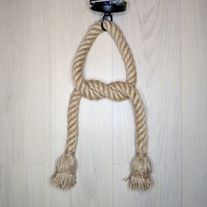 May include: A ceiling light fitting component featuring a thick, natural tan jute rope looped through a black metal circular ceiling canopy. The rope is tied in a central knot, with two hanging ends finished with frayed, tassel-like rope ends.