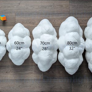 May include: Six white, cloud-shaped, textured decorative pillows arranged in increasing size from 12 inches to 36 inches. Each pillow is labeled with its corresponding width in centimeters and inches. A small, colorful cube sits near the smallest pillow.