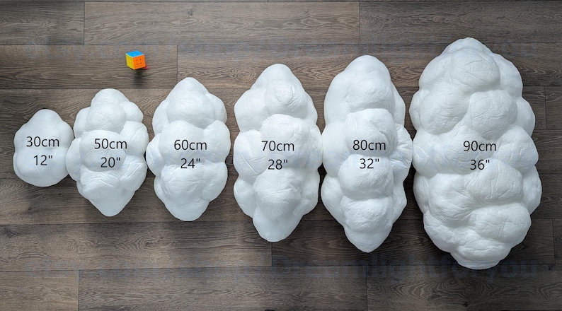 May include: Six white, cloud-shaped, textured decorative pillows arranged in increasing size from 12 inches to 36 inches. Each pillow is labeled with its corresponding width in centimeters and inches. A small, colorful cube sits near the smallest pillow.