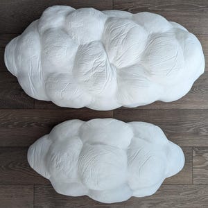 May include: Two white, cloud-shaped decorative objects with a textured, matte surface. The larger cloud is positioned above the smaller cloud. Both items feature a rounded, puffy, multi-lobed form with visible creases and indentations across the surface.