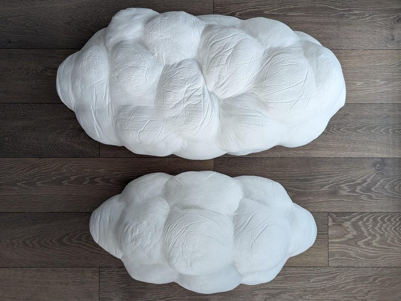 May include: Two white, cloud-shaped decorative objects with a textured, matte surface. The larger cloud is positioned above the smaller cloud. Both items feature a rounded, puffy, multi-lobed form with visible creases and indentations across the surface.