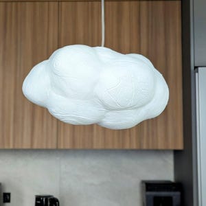 May include: A white pendant light fixture shaped like a fluffy cloud with a textured, uneven surface. The lamp is suspended from the ceiling by a thin white cord, featuring rounded, bulbous sections that create a soft, organic, and voluminous form.