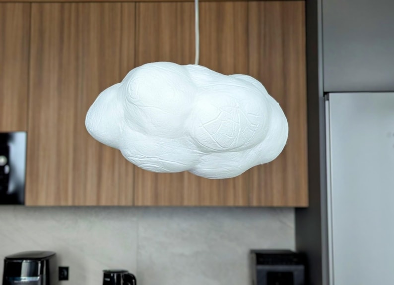 May include: A white pendant light fixture shaped like a fluffy cloud with a textured, uneven surface. The lamp is suspended from the ceiling by a thin white cord, featuring rounded, bulbous sections that create a soft, organic, and voluminous form.