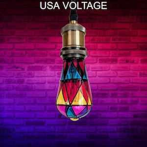 Puede incluir: Una bombilla de cristal de colores con una fijación de latón. La bombilla tiene forma de lágrima y presenta paneles de vidrio rojo, amarillo, azul y verde. La bombilla está colgada de una pared de ladrillo con un degradado de rosa y morado.