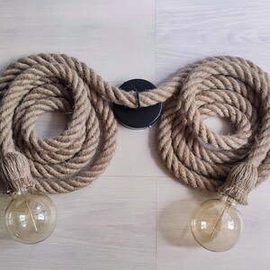 May include: A double light fitting featuring two thick, coiled natural tan jute ropes connected to a central black circular ceiling mount. Each rope end is finished with a matching jute-wrapped socket holding a clear, round Edison-style light bulb.