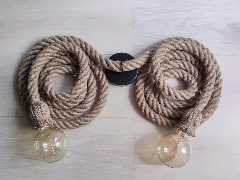 May include: A double light fitting featuring two thick, coiled natural tan jute ropes connected to a central black circular ceiling mount. Each rope end is finished with a matching jute-wrapped socket holding a clear, round Edison-style light bulb.