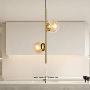 Op de afbeelding: Moderne hanglamp met twee bolvormige amberkleurige glazen kappen. Het licht hangt aan een goudkleurige metalen staaf. De armatuur hangt boven een aanrecht, met een witte muur op de achtergrond.