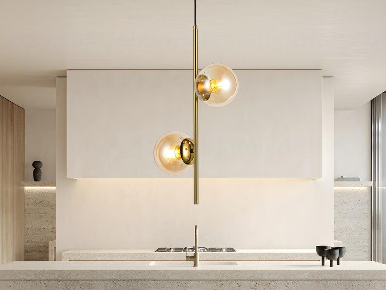 Op de afbeelding: Moderne hanglamp met twee bolvormige amberkleurige glazen kappen. Het licht hangt aan een goudkleurige metalen staaf. De armatuur hangt boven een aanrecht, met een witte muur op de achtergrond.
