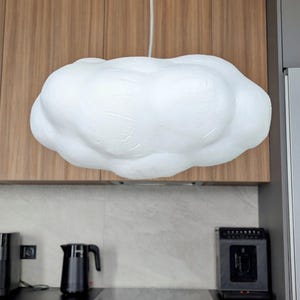 May include: A white, cloud-shaped pendant light fixture with a textured, matte surface. The lamp is suspended from the ceiling by a single white cord, featuring a rounded, organic form that mimics the appearance of a fluffy cumulus cloud.