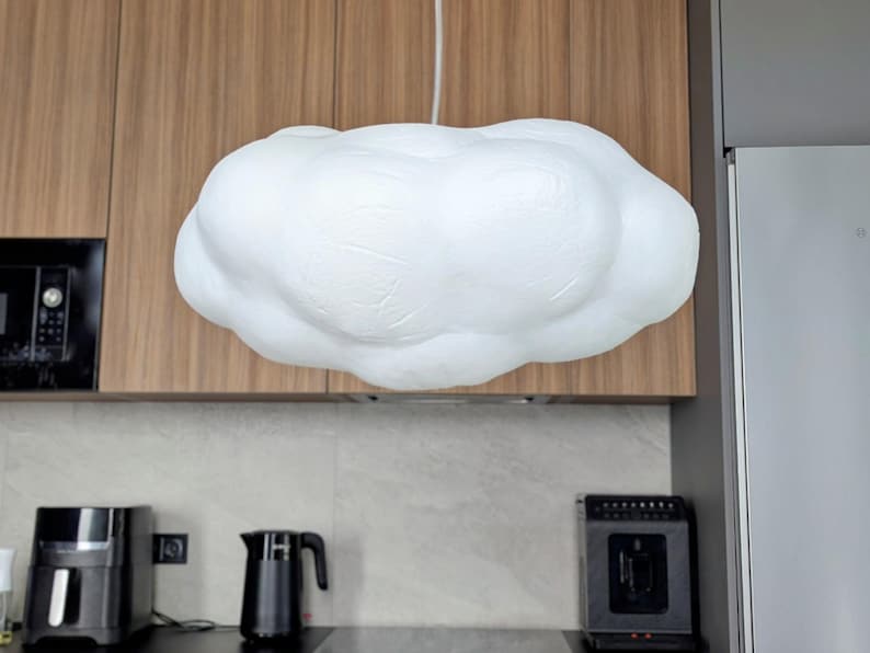 May include: A white, cloud-shaped pendant light fixture with a textured, matte surface. The lamp is suspended from the ceiling by a single white cord, featuring a rounded, organic form that mimics the appearance of a fluffy cumulus cloud.