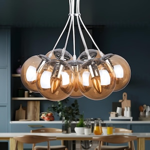 May include: A modern chandelier with seven spherical amber-colored glass shades. The light fixture has chrome accents and white cords. The chandelier hangs above a white table in a kitchen setting.