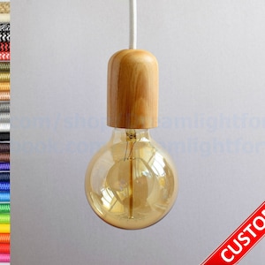 Puede incluir: Una lámpara colgante de madera con una bombilla de vidrio transparente. La lámpara tiene un acabado de madera natural y un diseño sencillo. La bombilla es una bombilla Edison estándar con un brillo blanco cálido. La palabra "CUSTOMIZED" está impresa en rojo sobre un fondo blanco en la esquina inferior derecha de la imagen.
