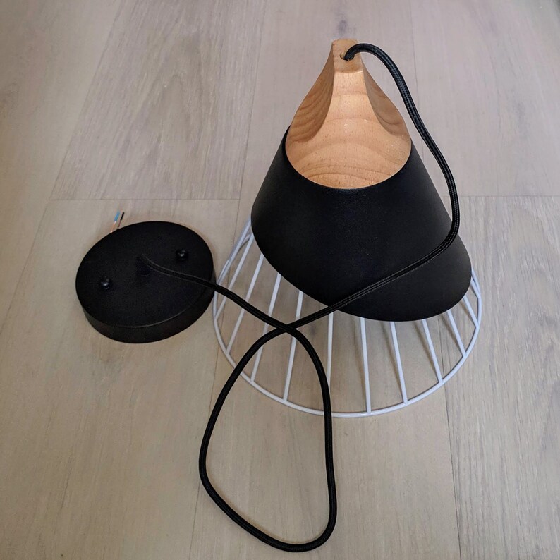 Pu&ograve; includere: Una lampada a sospensione con una parte superiore in legno naturale, un paralume conico nero opaco e una base a gabbia metallica bianca. Il dispositivo include un rosone circolare nero e un cavo elettrico rivestito in tessuto nero.