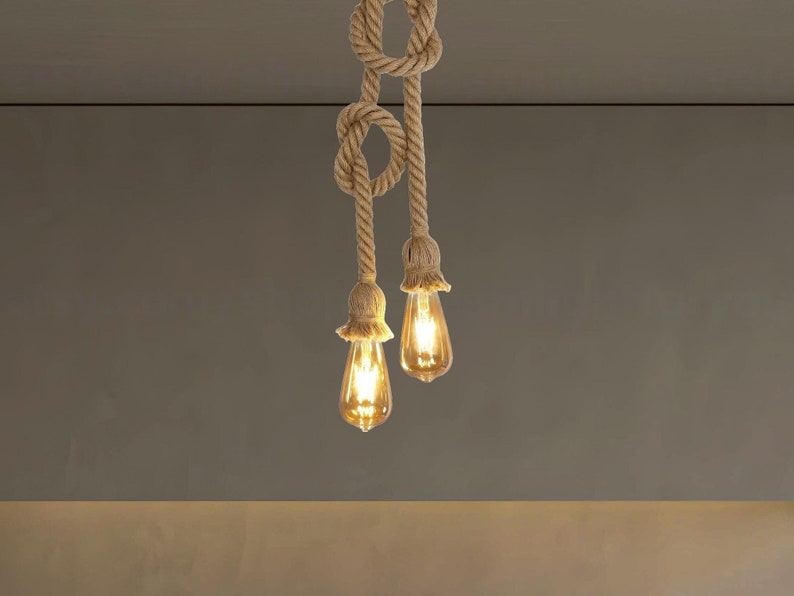 May include: A dual-pendant light fixture featuring two thick, tan-coloured twisted hemp ropes tied into decorative knots. Each rope terminates in a flared, frayed rope socket housing a glowing, amber-tinted Edison-style light bulb.