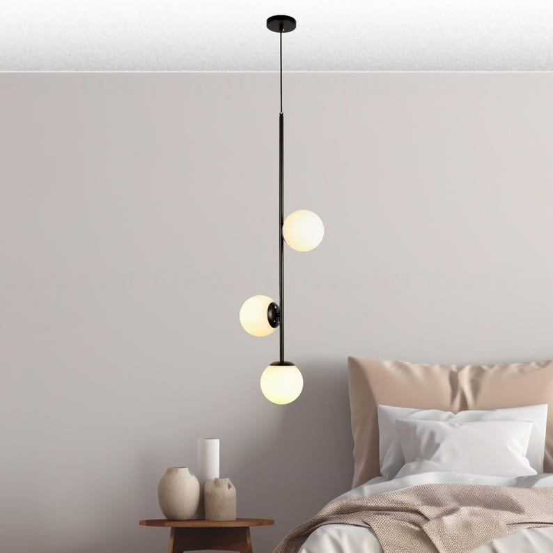Op de afbeelding: Een moderne zwarte hanglamp met drie bolvormige, matglazen kappen. Het licht hangt verticaal, met de kappen verdeeld over een slanke zwarte staaf. De armatuur is opgehangen aan een zwarte plafondbevestiging.