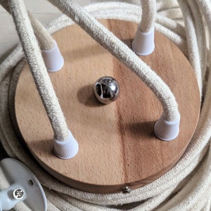 May include: A wooden ceiling rose with three white cord covers and a silver metal ball in the center. The rose is surrounded by a coiled length of white fabric-covered electrical cord.