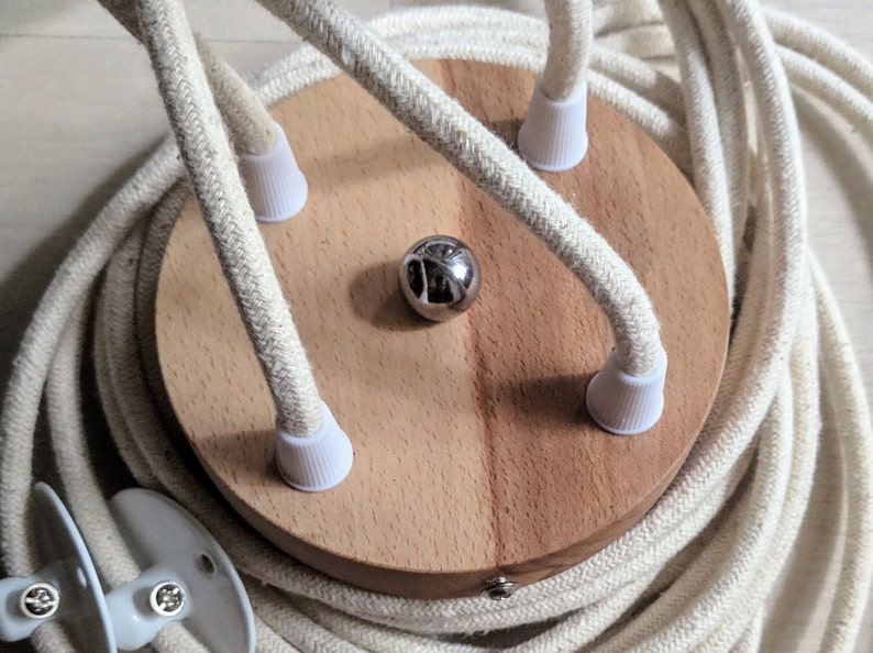 May include: A wooden ceiling rose with three white cord covers and a silver metal ball in the center. The rose is surrounded by a coiled length of white fabric-covered electrical cord.