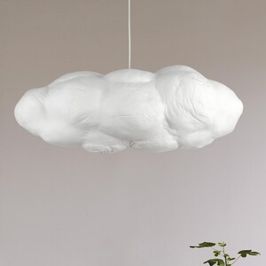 May include: A white pendant light fixture shaped like a fluffy cloud. The surface has a textured, matte appearance with rounded, organic contours. It is suspended from the ceiling by a thin, vertical white cord centered at the top of the form.