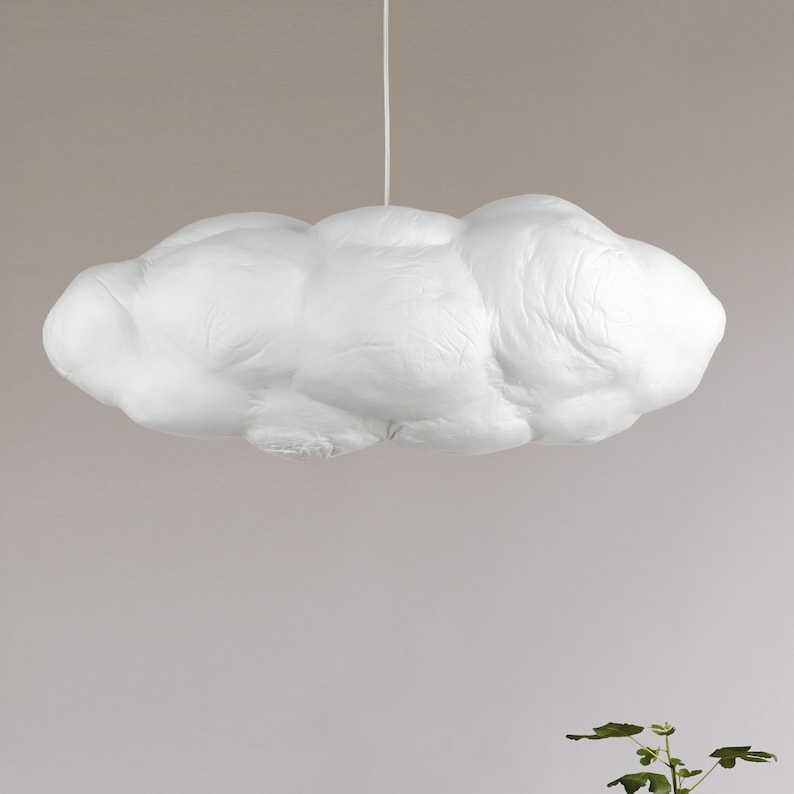 May include: A white pendant light fixture shaped like a fluffy cloud. The surface has a textured, matte appearance with rounded, organic contours. It is suspended from the ceiling by a thin, vertical white cord centered at the top of the form.