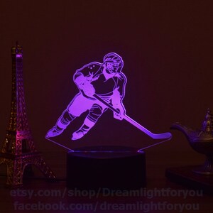 May include: A purple 3D printed night light depicting a hockey player skating with a stick. The light is on a black base and is illuminated. The image also shows a decorative lamp and a miniature Eiffel Tower.