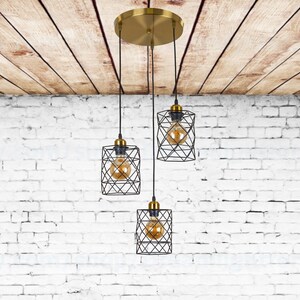May include: Three black wire cage pendant lights with gold accents hang from a gold ceiling fixture against a white brick wall.