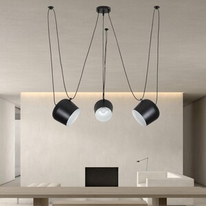 May include: Three black pendant lights with white interior shades hang from the ceiling on long cords. The lights are suspended over a wooden table in a modern living room.