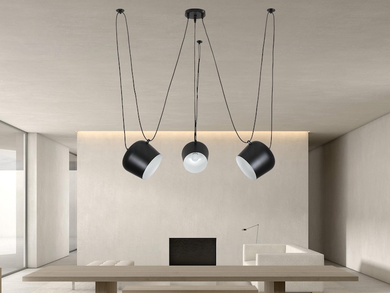 May include: Three black pendant lights with white interior shades hang from the ceiling on long cords. The lights are suspended over a wooden table in a modern living room.