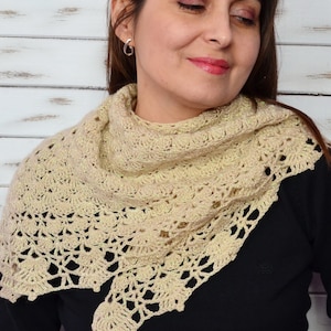 May include: A beige crocheted shawl with a delicate, openwork design. The shawl is draped over the shoulders, showcasing a textured, lacy pattern. The yarn has a subtle shimmer, adding a touch of elegance to the handmade accessory.