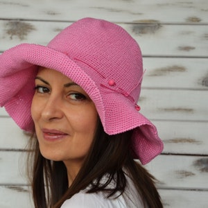 A pink crocheted sun hat with a wide brim and pink beads. The hat is worn by a person with long brown hair.