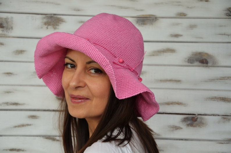 A pink crocheted sun hat with a wide brim and pink beads. The hat is worn by a person with long brown hair.