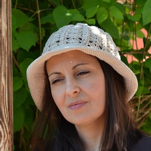 May include: A woman wearing a beige crocheted sun hat with a wide brim. The hat has a delicate openwork pattern. The woman has long brown hair and is looking down.