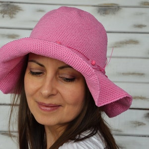 A pink crochet sun hat with a wide brim and a decorative bow. The hat has a textured surface and is adorned with small pink beads.