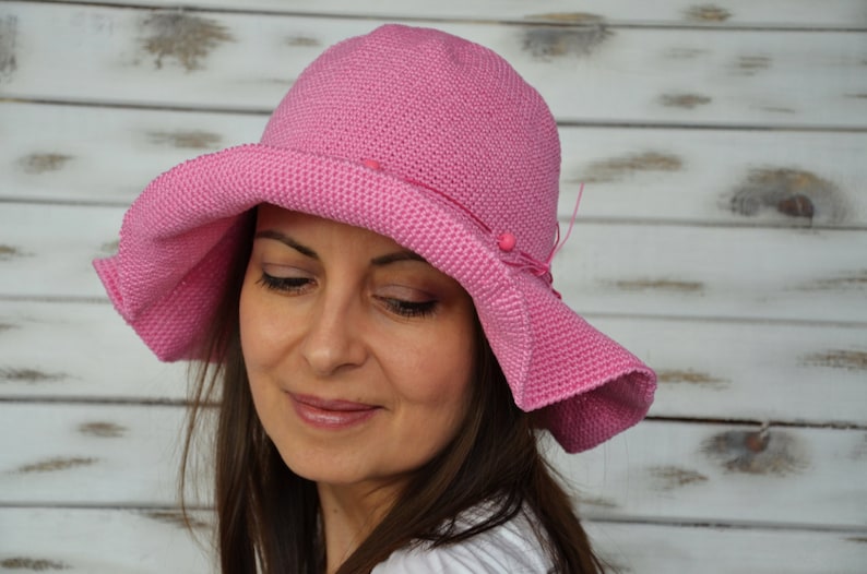A pink crochet sun hat with a wide brim and a decorative bow. The hat has a textured surface and is adorned with small pink beads.