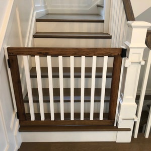 May include: A white and brown wooden baby gate with vertical slats, installed at the top of a staircase. The gate is attached to the wall and the railing with metal brackets.