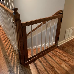 May include: A wooden baby gate with white spindles installed at the top of a staircase. The gate is made of stained wood and has a simple design.