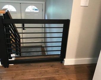 May include: Black metal baby gate with horizontal bars, installed at the top of a staircase. The gate is on wheels and has a rectangular frame. The stairs have a wooden handrail and the floor is made of wood.