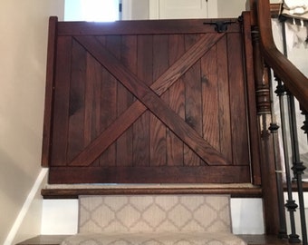 May include: A dark brown wooden gate with an X-shaped design, installed at the top of a carpeted staircase. The gate is closed and appears to be a safety barrier for children or pets.
