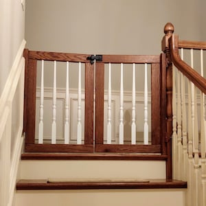 May include: A wooden baby gate with white spindles, installed at the top of a staircase. The gate is closed and has a black latch.