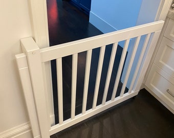 May include: White wooden baby gate with vertical slats, installed in a doorway with white trim and dark hardwood floors.