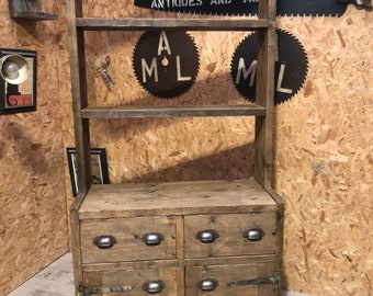 May include: A rustic wooden shelf unit with two drawers and two cabinet doors. The unit has a distressed brown finish and features metal hardware. The shelf unit is against a wood panel background with various signs and decorations.