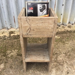 May include: A rustic wooden record stand with two shelves. The top shelf has a vinyl record with the title "Lucille B.B. King" on it. The stand is made of reclaimed wood and has a natural finish.