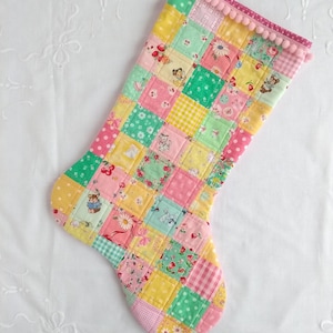 May include: A patchwork Christmas stocking with a pink and white pom-pom trim. The stocking is made of various fabrics in shades of pink, green, yellow, and white, with a variety of small floral and animal prints.