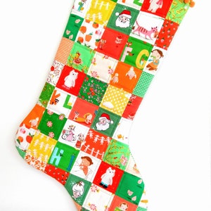 Könnte beinhalten: Ein grüner und orangefarbener Patchwork-Weihnachtsstrumpf mit weißem Bommelbesatz. Der Strumpf ist mit verschiedenen Bildern verziert, darunter der Weihnachtsmann, ein Weihnachtsbaum und Buchstaben des Alphabets.