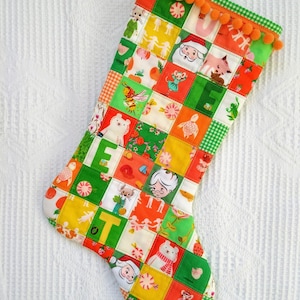 May include: A patchwork Christmas stocking in green, orange and white. The stocking is decorated with various festive images, including Santa Claus, a polar bear and a bird. The stocking has a pom-pom trim and the word "JOY" is spelled out in the design.